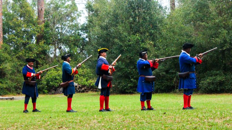 A line of 5 reenactors wearing blue and red historic costume prepare to fire muskets. 